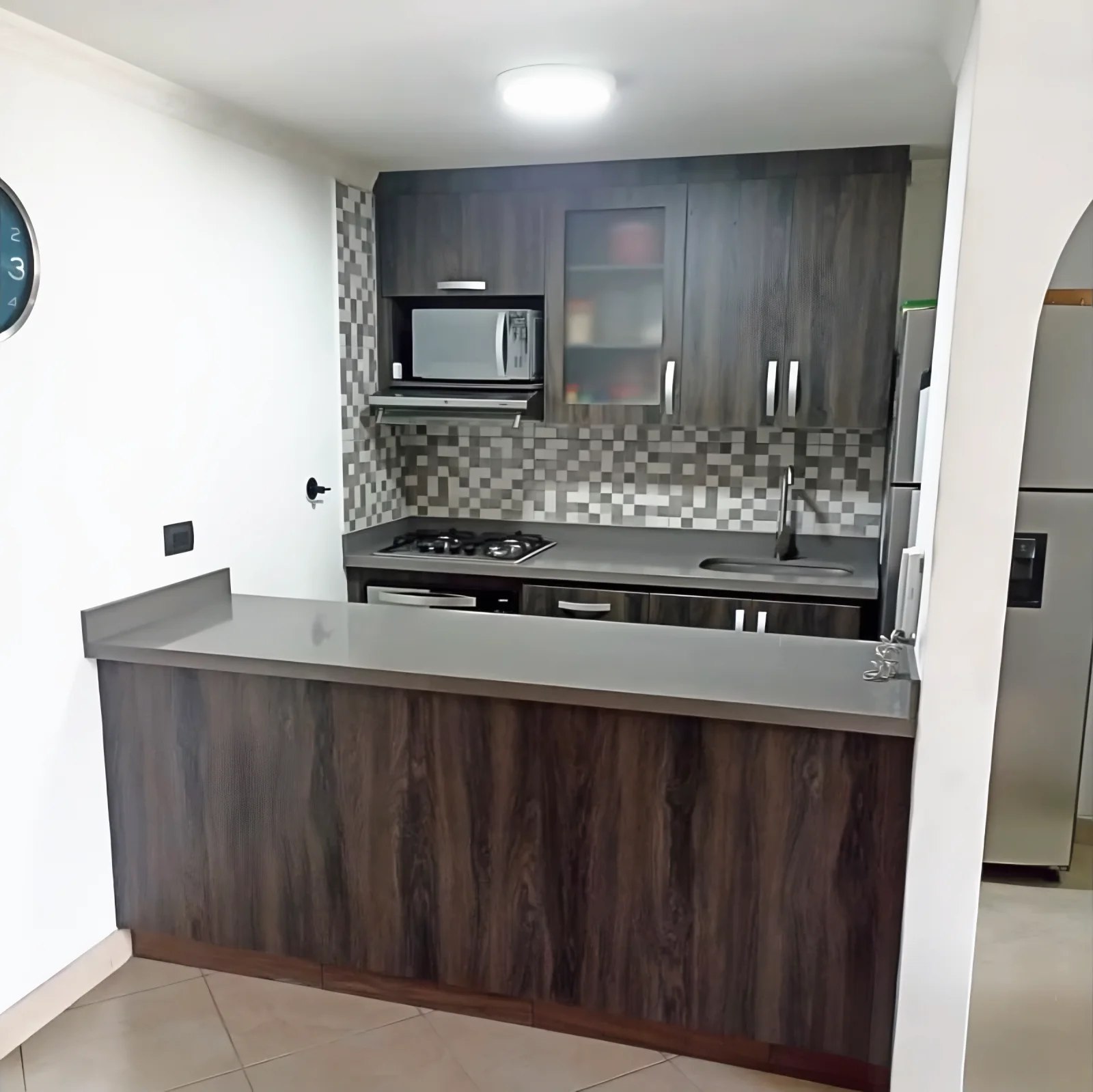 Cocina con barra forrada en madera oscura, mesón gris, alacenas oscuras con vitrina y microondas empotrado